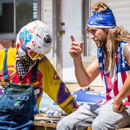 Two people in colorful attire, one wearing Blue Ribbon Beer Goggles and a helmet, outdoors with a relaxed vibe.