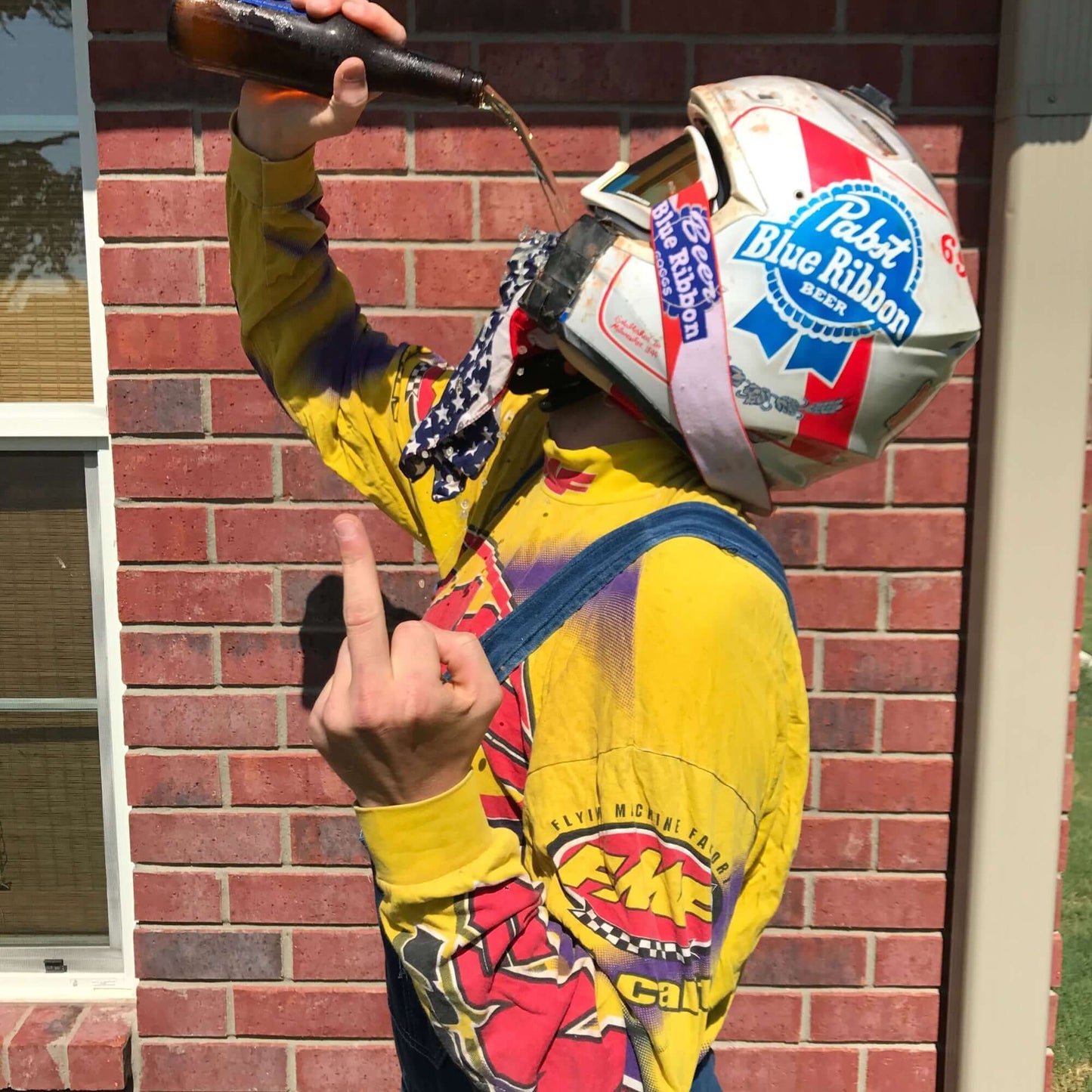 Person wearing Blue Ribbon Goggles and colorful gear pouring beer into their helmet in front of a brick wall.