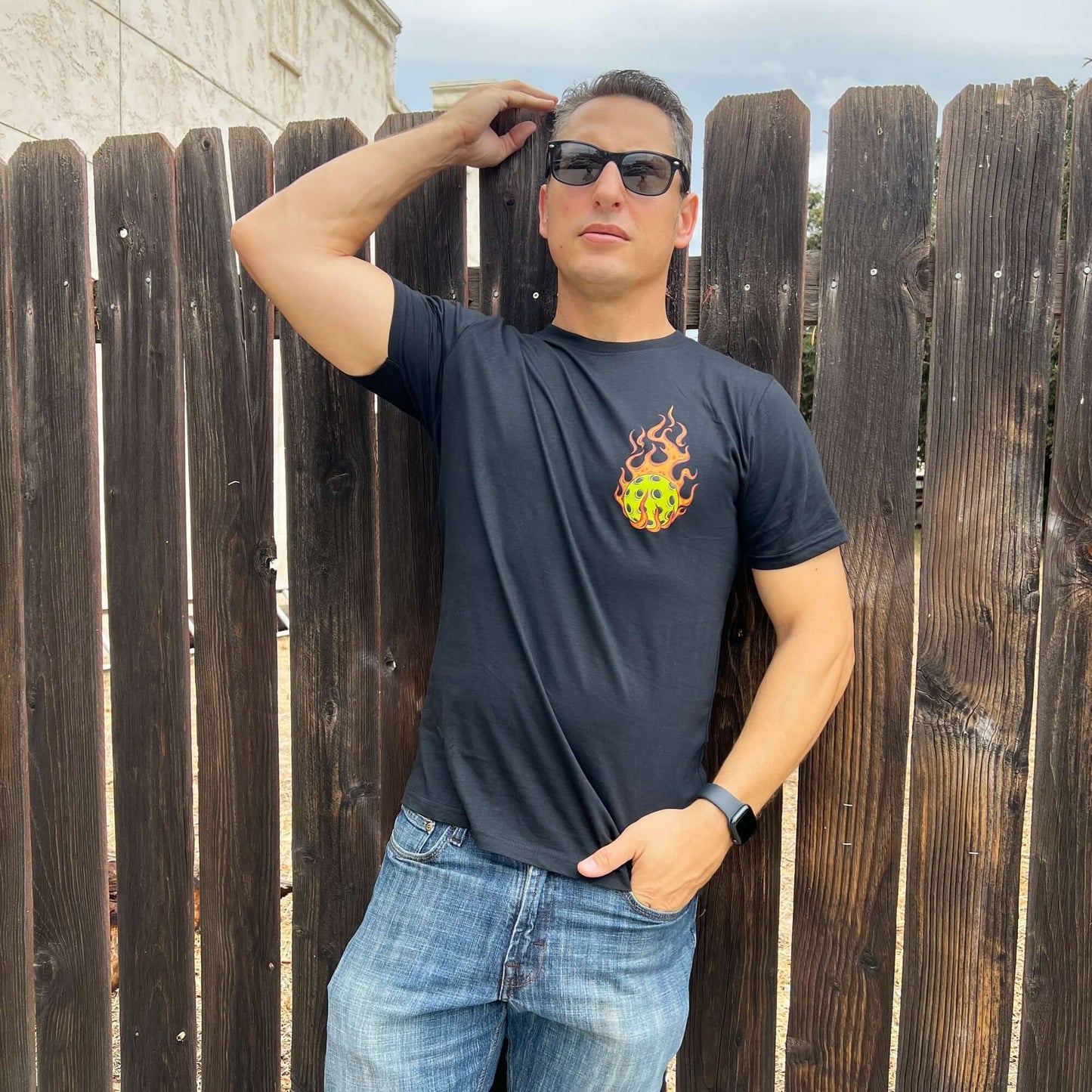 Man wearing Flaming Pickleball Black T-Shirt with fiery design, posing confidently against a wooden fence.