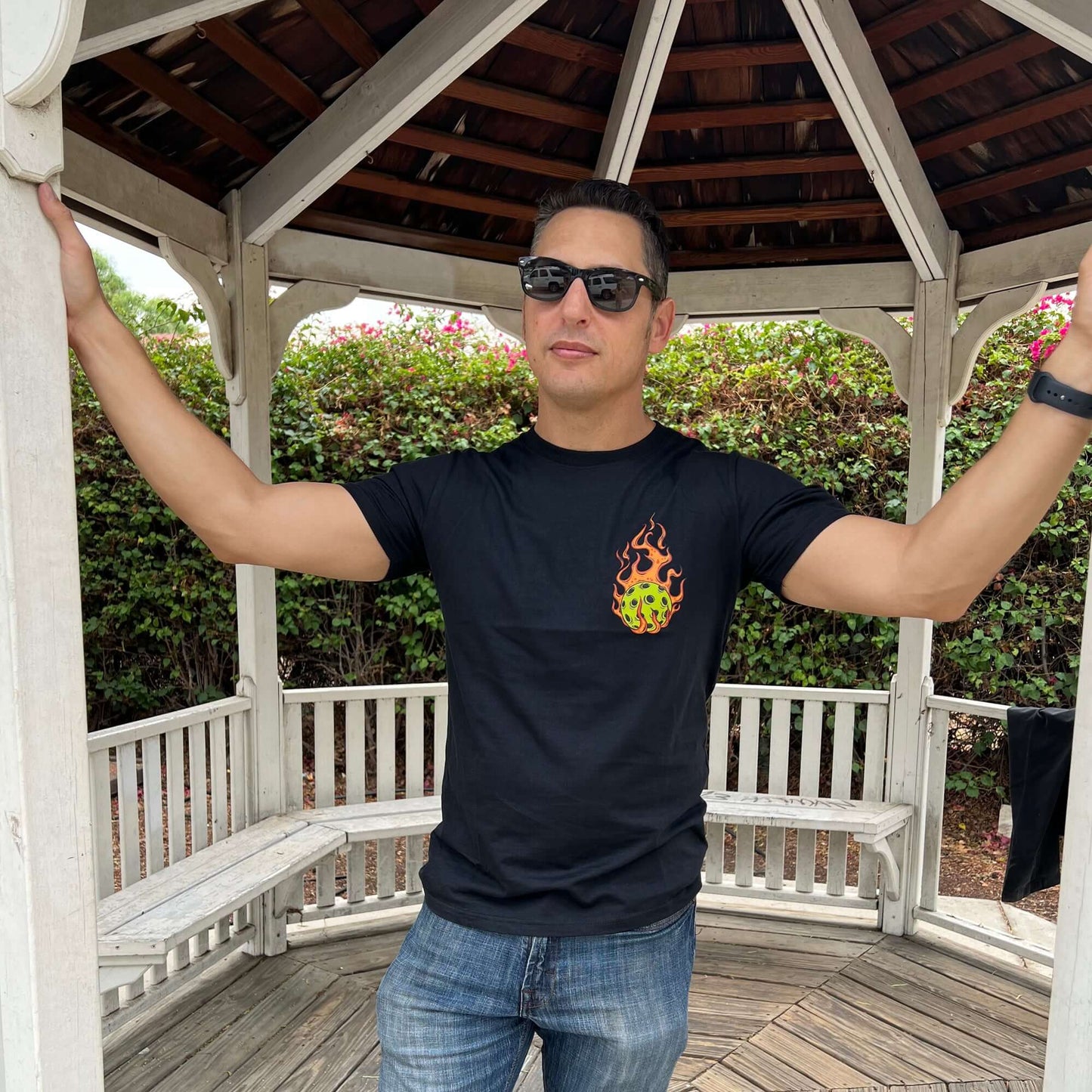 Man wearing a Flaming Pickleball Black T-Shirt standing in a gazebo, exuding confidence and ready to dominate the competition.