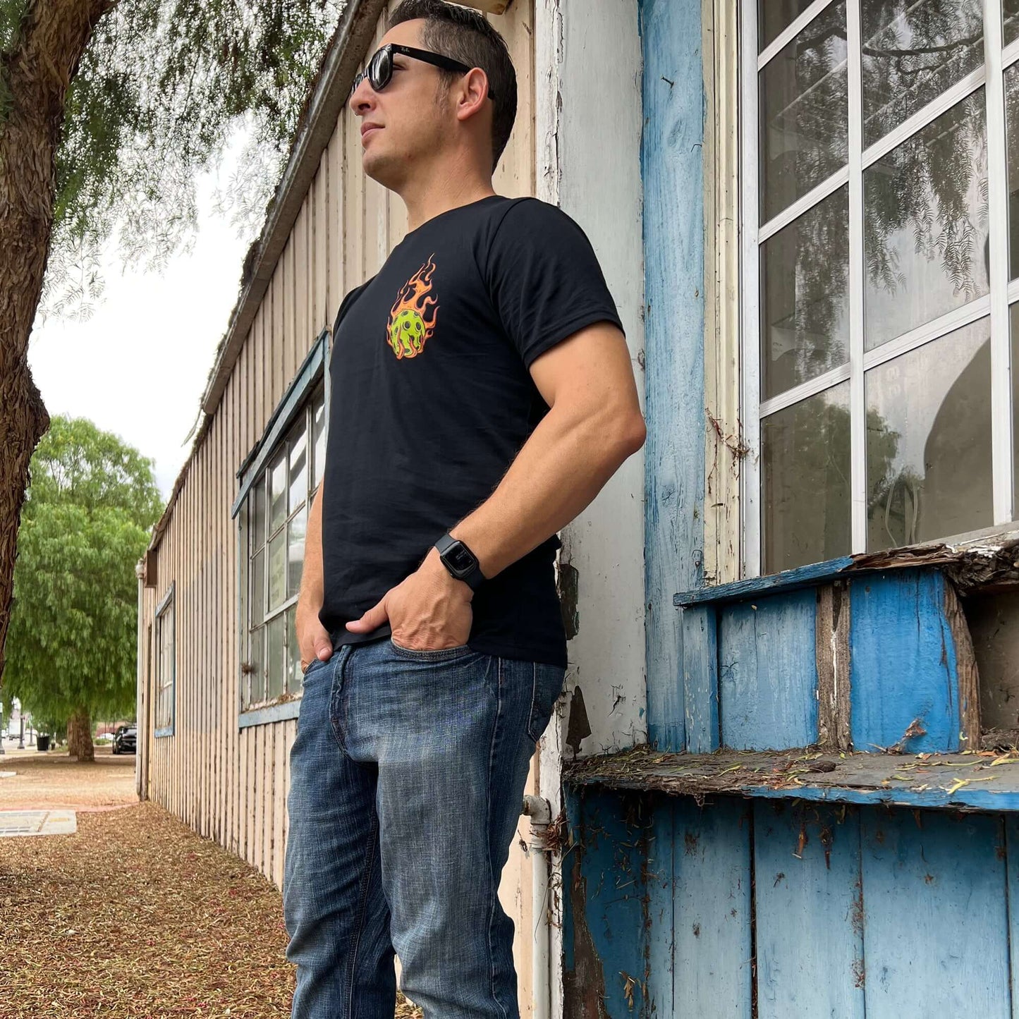 Man wearing Flaming Pickleball Black T-Shirt standing by rustic building, showcasing fiery design for competitive inspiration.
