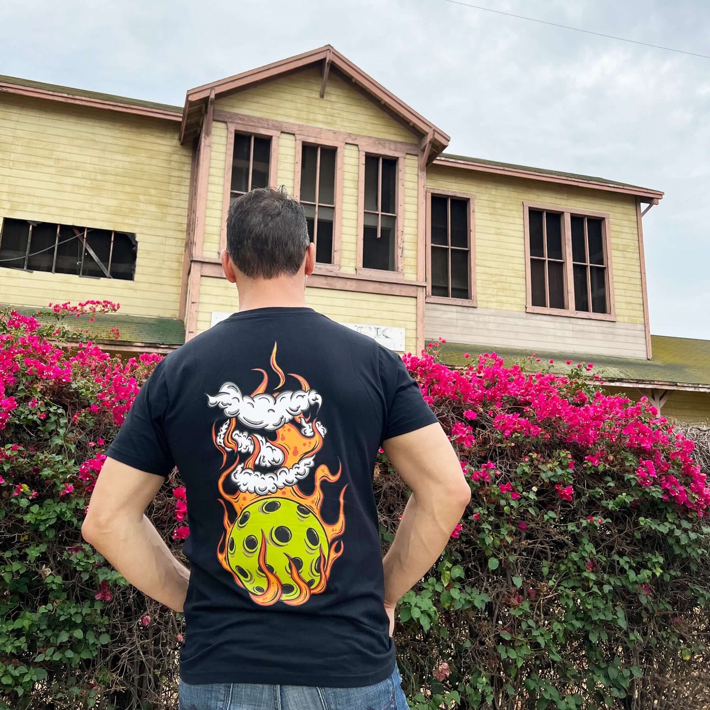 Man wearing Flaming Pickleball Black T-Shirt with fiery design, standing in front of an old building and pink flowers.