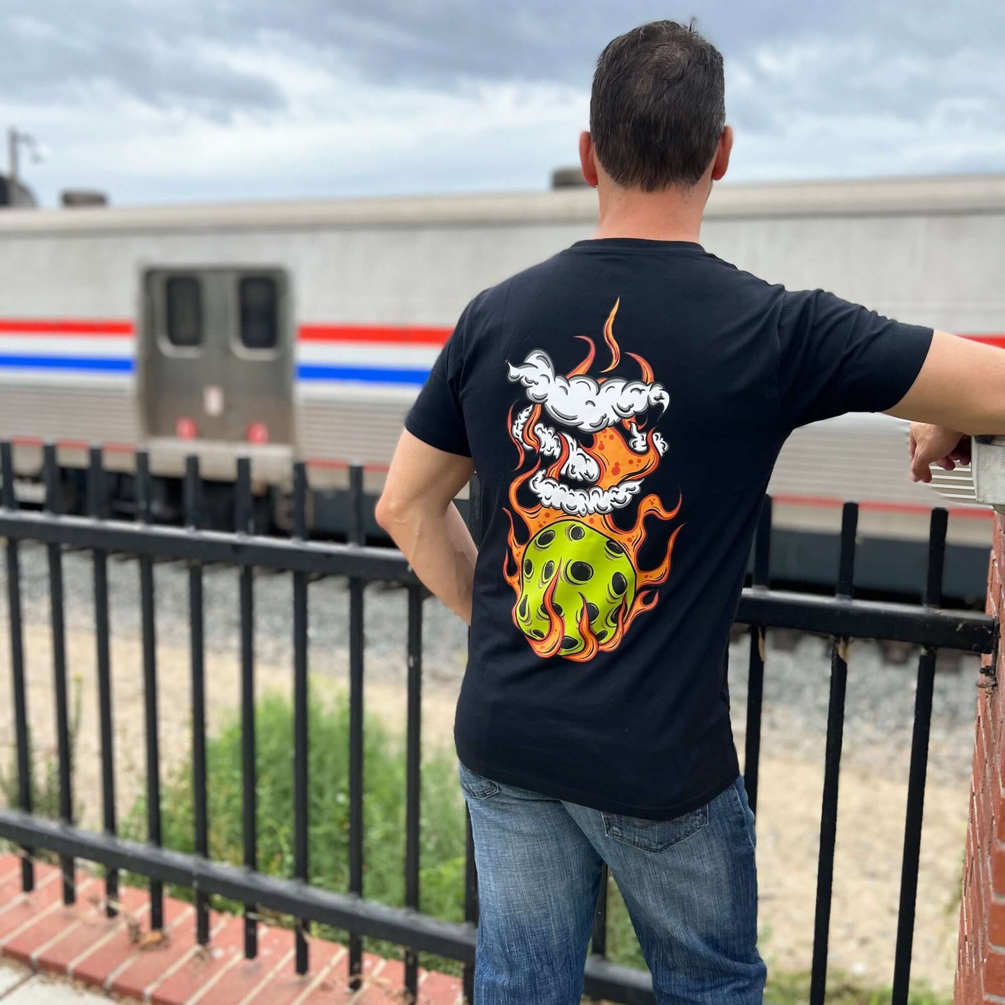 Man wearing black flaming pickleball T-shirt, standing near a train, showcasing the bold graphic design on the back.