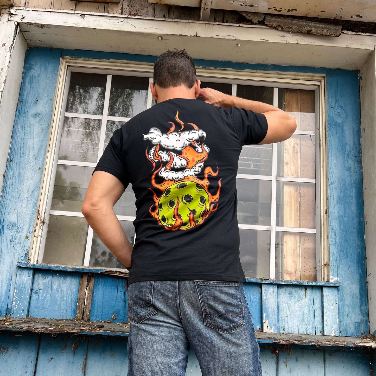 Man wearing a Flaming Pickleball Black T-Shirt with bold fiery pickleball design on the back, standing outdoors.