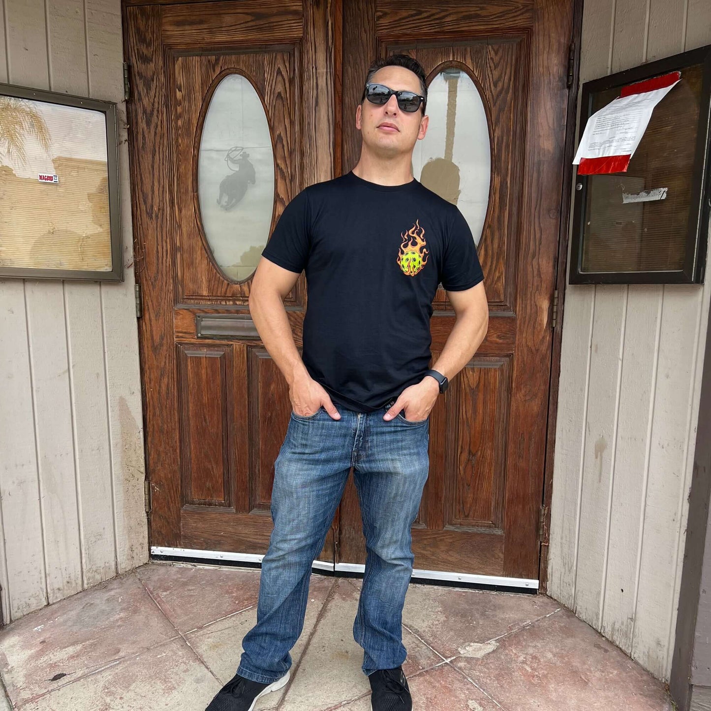 Man wearing a Flaming Pickleball Black T-Shirt stands confidently in front of rustic wooden doors.