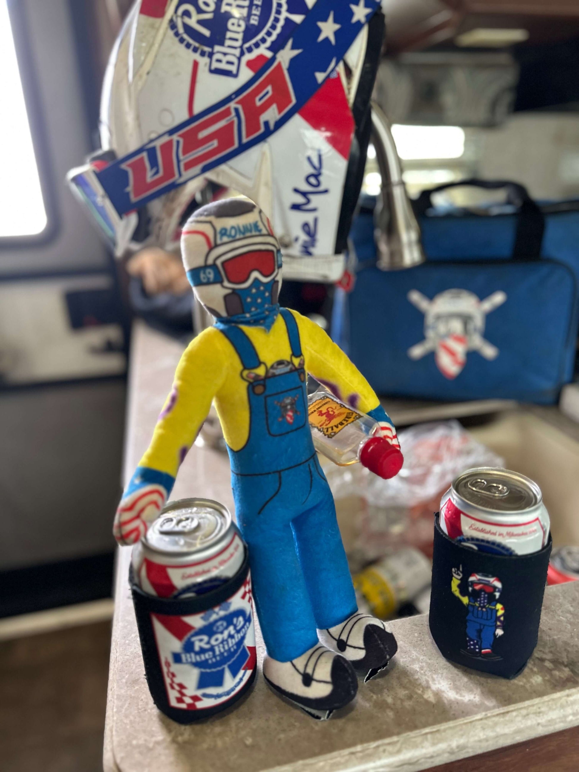 Lil Ronnie plush toy in denim overalls with two PBR cans, standing on a counter with USA-themed decor in the background.