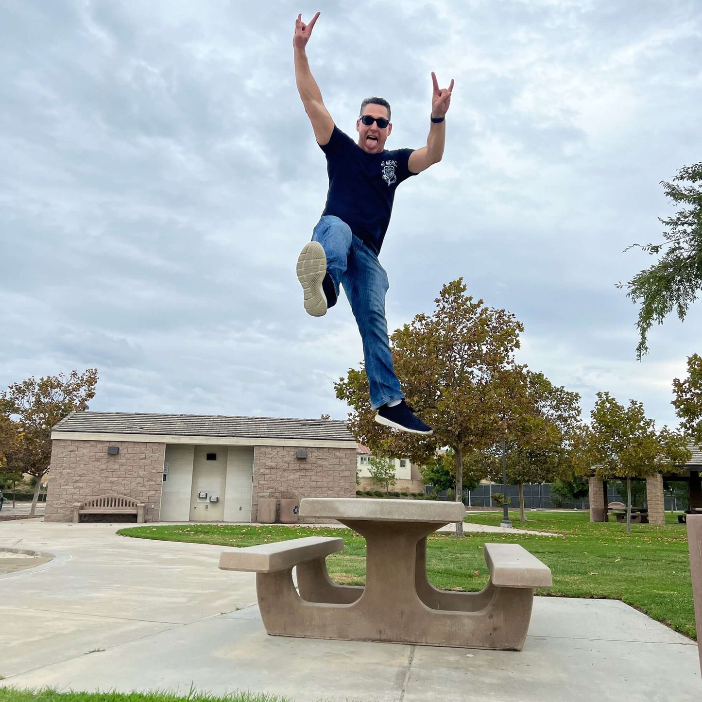 Man jumping off picnic table in park, wearing sunglasses and a black T-shirt, capturing a moment of joy and energy against a cloudy sky.