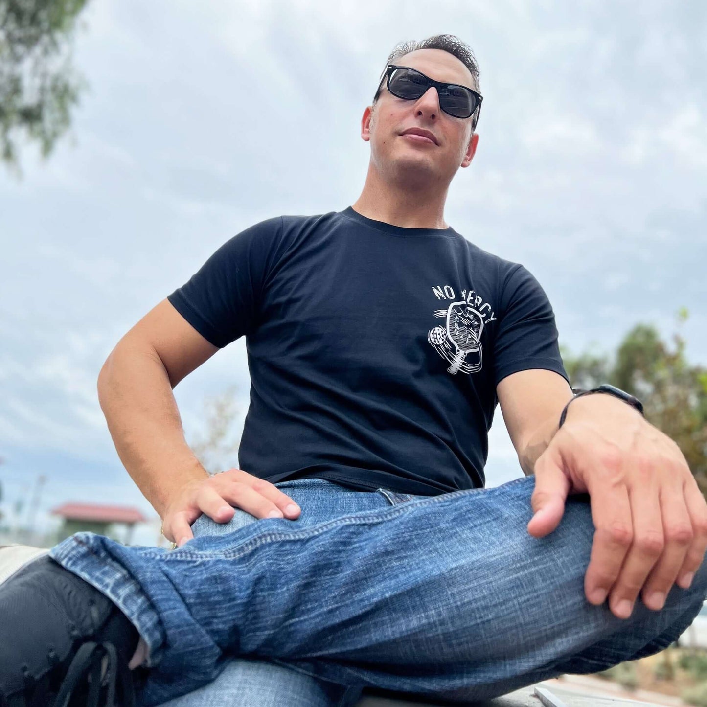 Man wearing a No Mercy Pickleball black T-shirt and sunglasses, sitting outdoors with a confident pose.