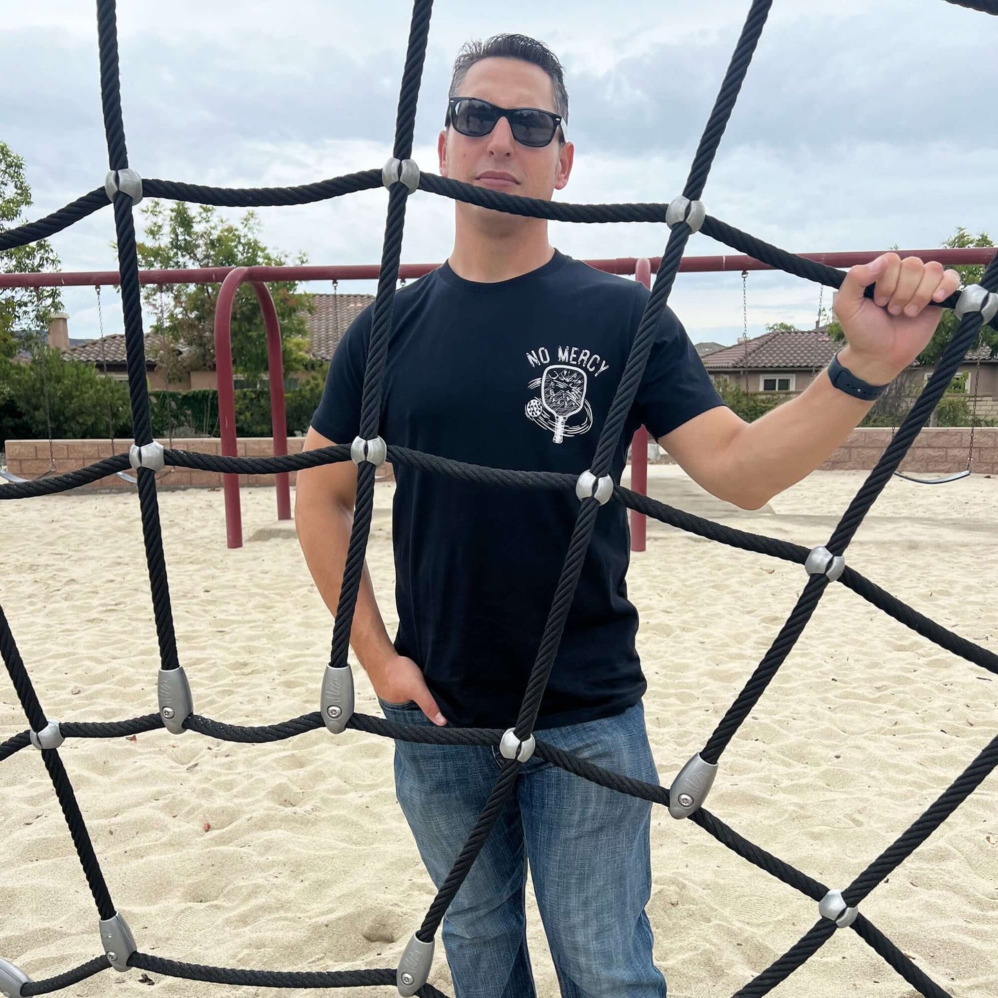 Man wearing No Mercy Pickleball Black T-Shirt standing behind a playground net outdoors.