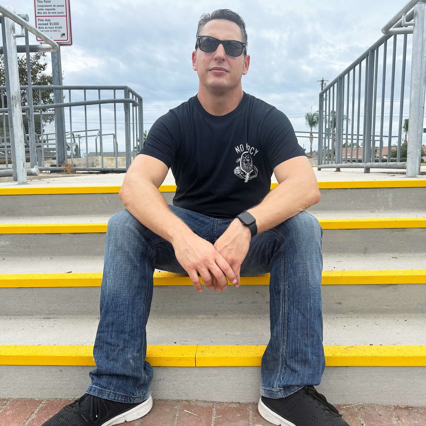 Man in sunglasses sitting on steps wearing No Mercy Pickleball Black T-Shirt, embodying aggressive and strategic spirit.