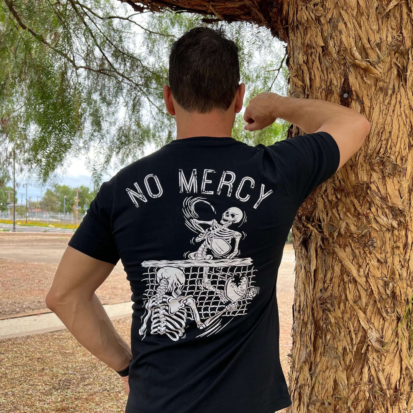 Man wearing No Mercy Pickleball black T-shirt with skeleton graphic, standing by tree in park.