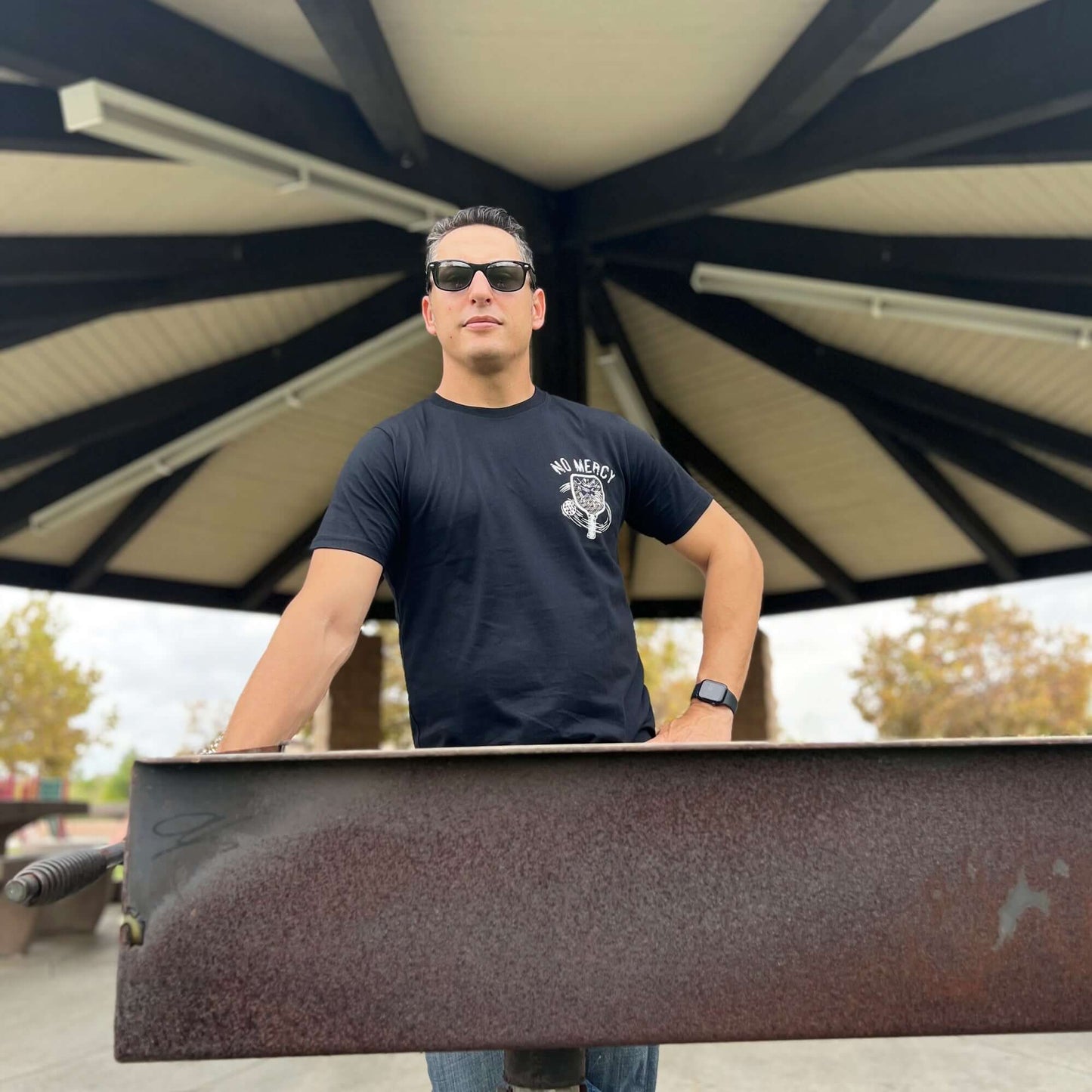 Person wearing black No Mercy Pickleball T-shirt standing confidently under a pavilion, embodying aggressive pickleball spirit.