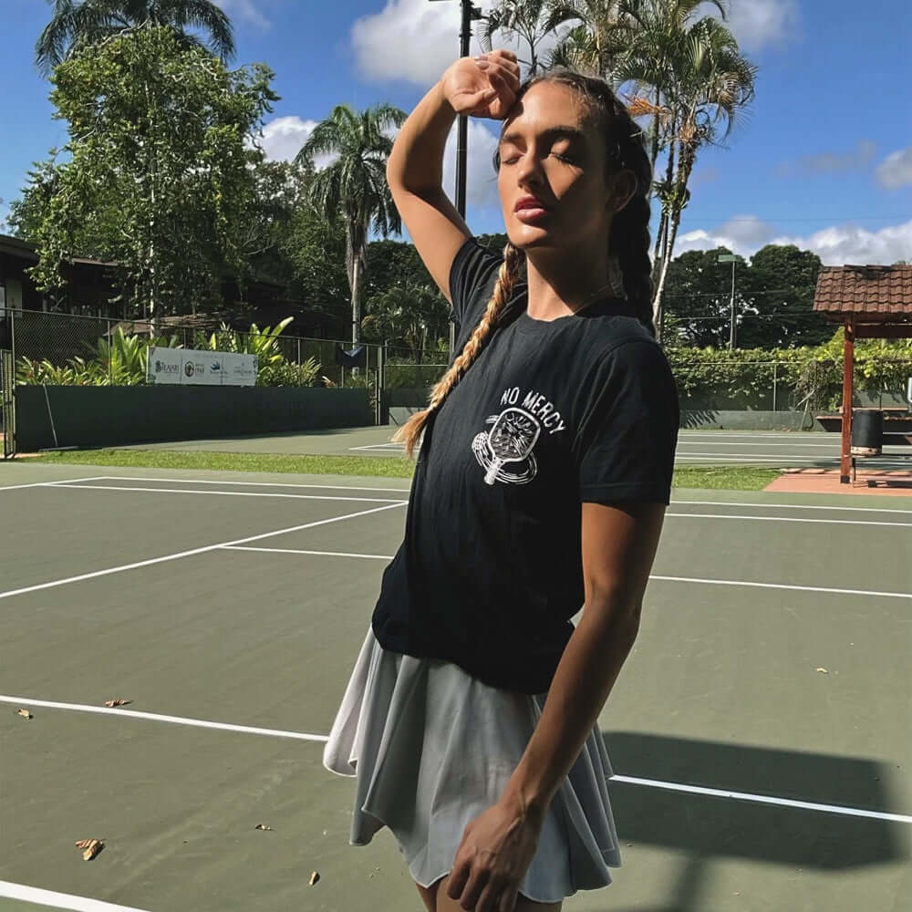 Woman wearing No Mercy Pickleball black T-shirt on a tennis court under sunny sky.