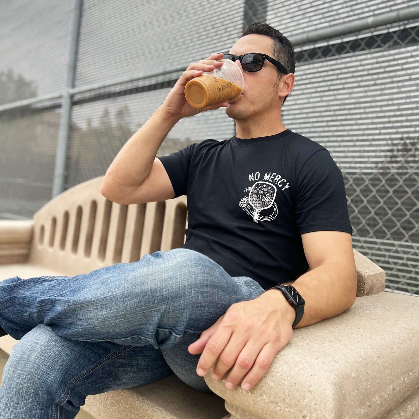 Person wearing No Mercy Pickleball Black T-Shirt sitting on a bench drinking coffee.