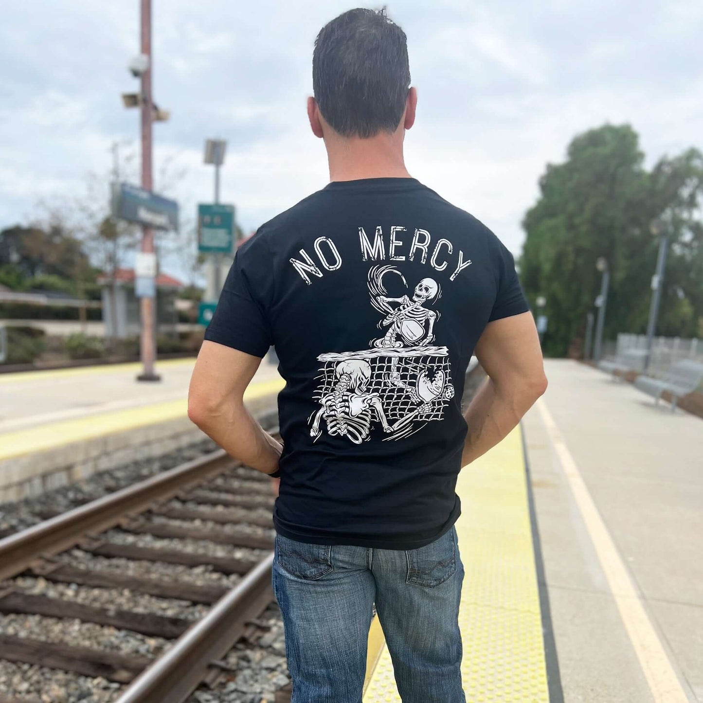 Man wearing No Mercy Pickleball black t-shirt with skeleton design standing on train platform.