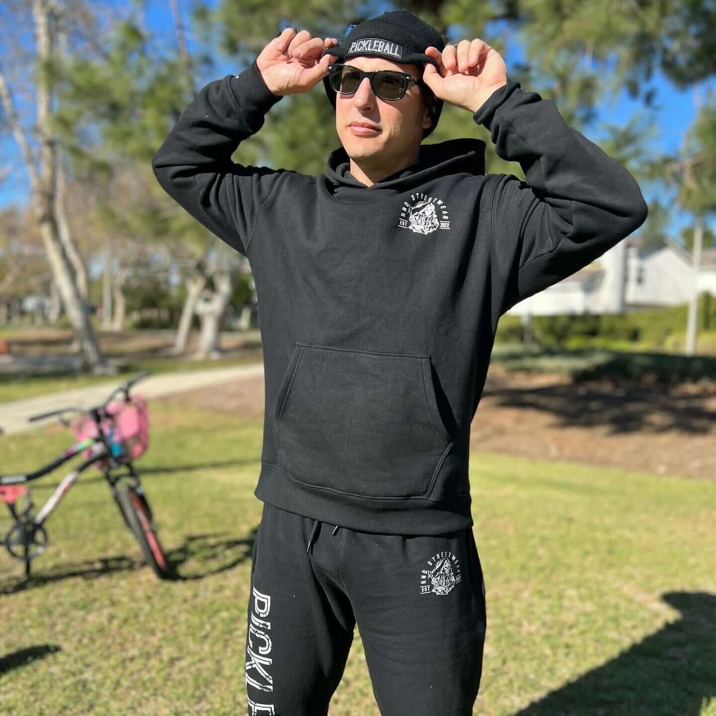 Person wearing a black Pickleball beanie, hoodie, and sweatpants outdoors.