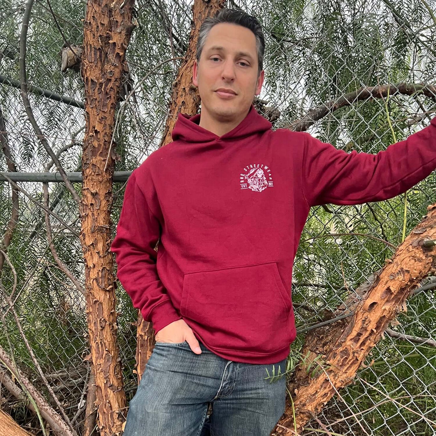 Man wearing a maroon hoodie with RND Streetwear logo, standing outdoors by trees and chain-link fence.