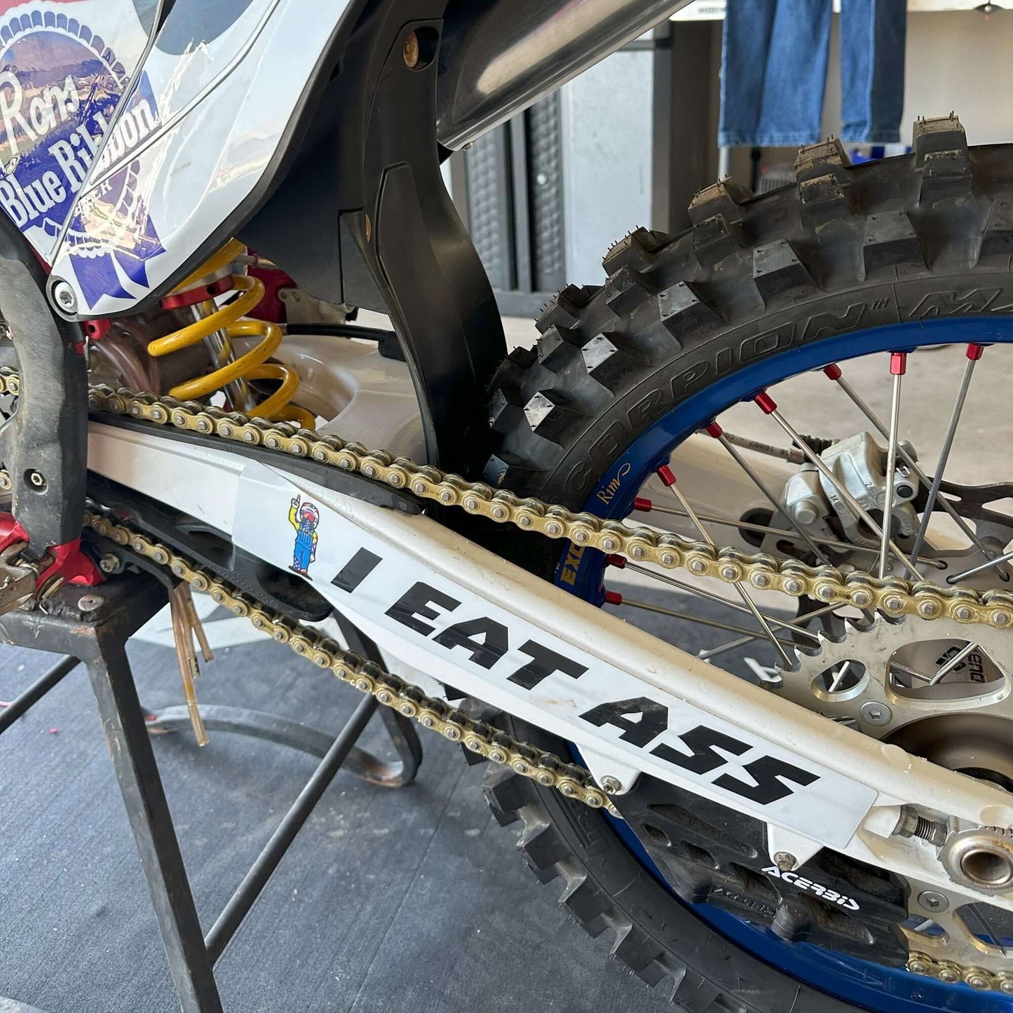 Swingarm dirt bike with "I EAT ASS" decal showing bold design and PBR branding in garage setting.