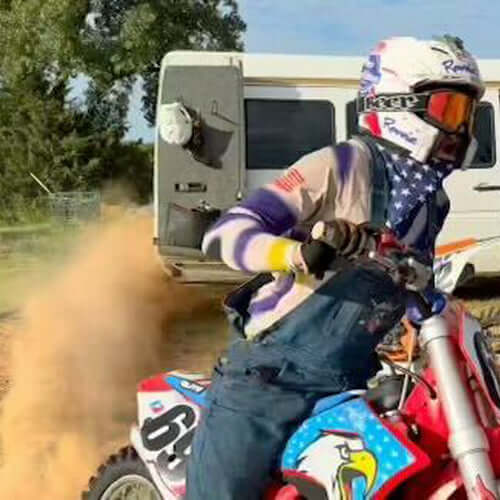 Motorcyclist in patriotic gear riding a dirt bike, kicking up dust near a camper in an outdoor setting.