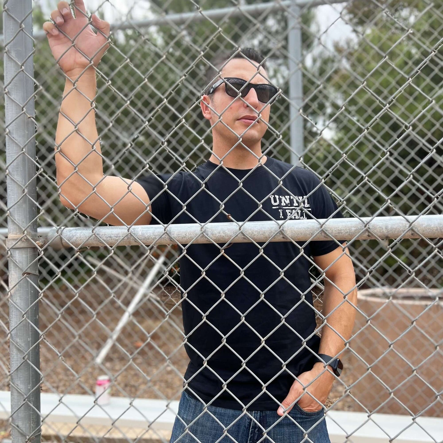 Man wearing black "Until I Fall" pickleball t-shirt standing by chain-link fence, embodying resilience and persistence.