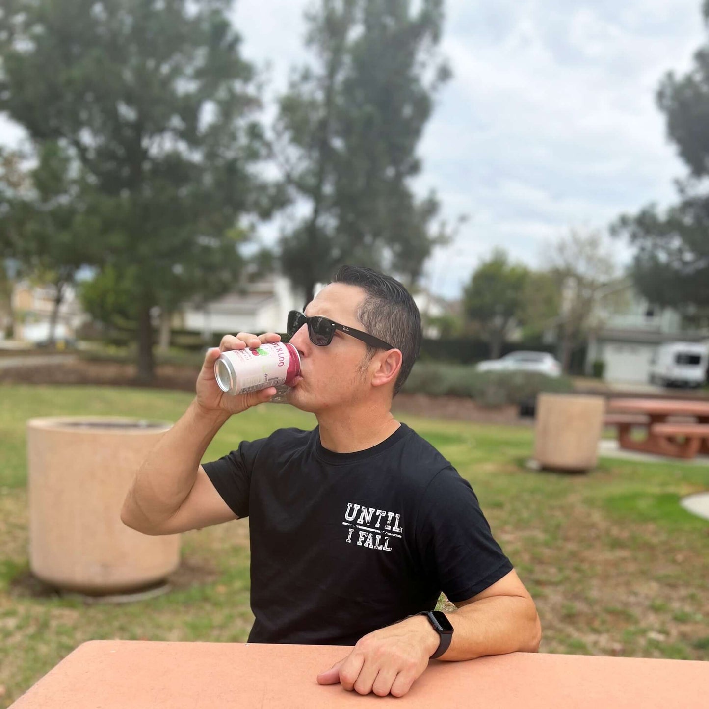 Person wearing an "Until I Fall" pickleball t-shirt drinks from a can outdoors at a park table.