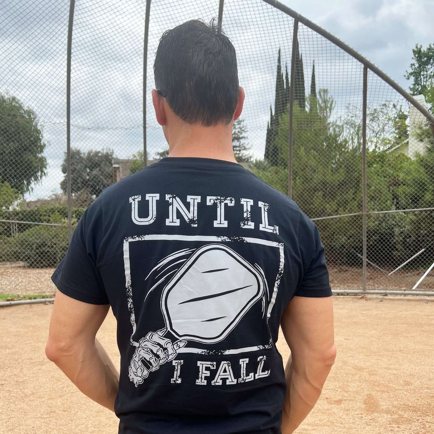 Man wearing the "Until I Fall" black pickleball t-shirt with paddle graphic, standing on a sports field.
