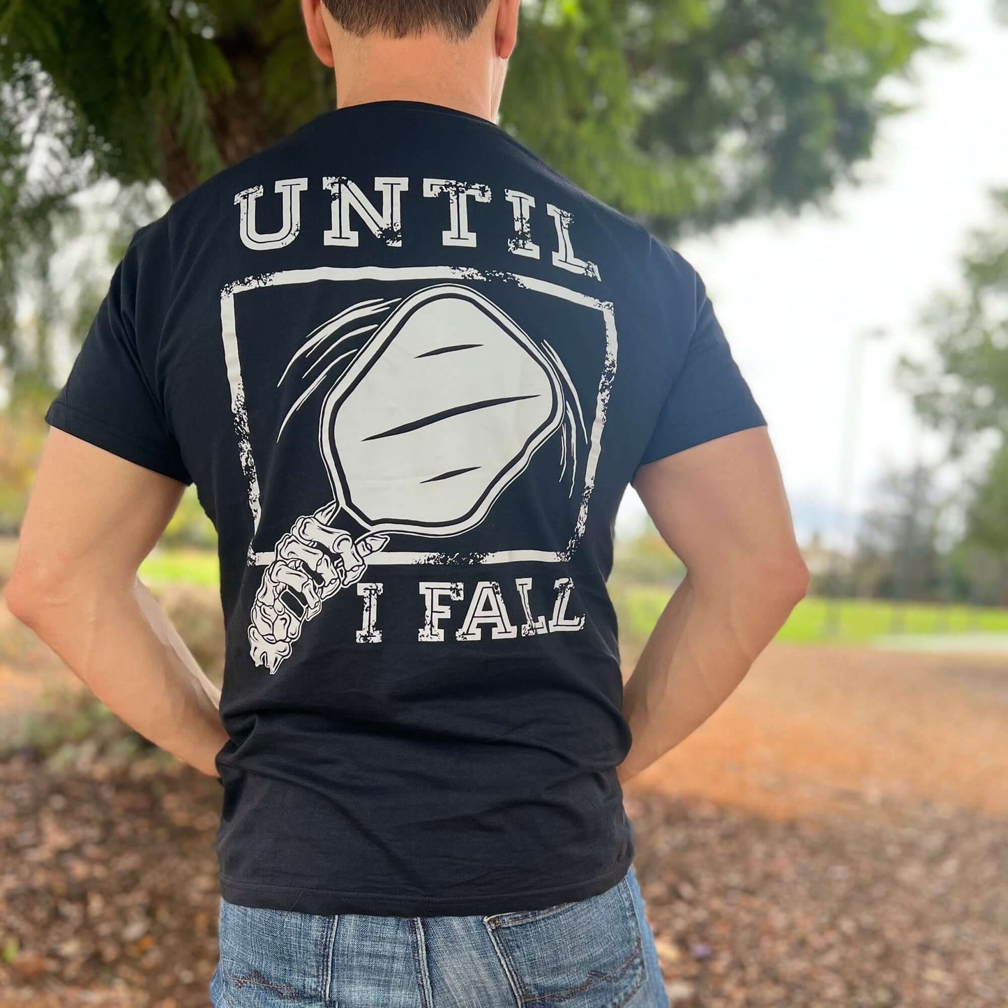 Man wearing a black "Until I Fall" pickleball t-shirt featuring a paddle design, standing on a tree-lined path.