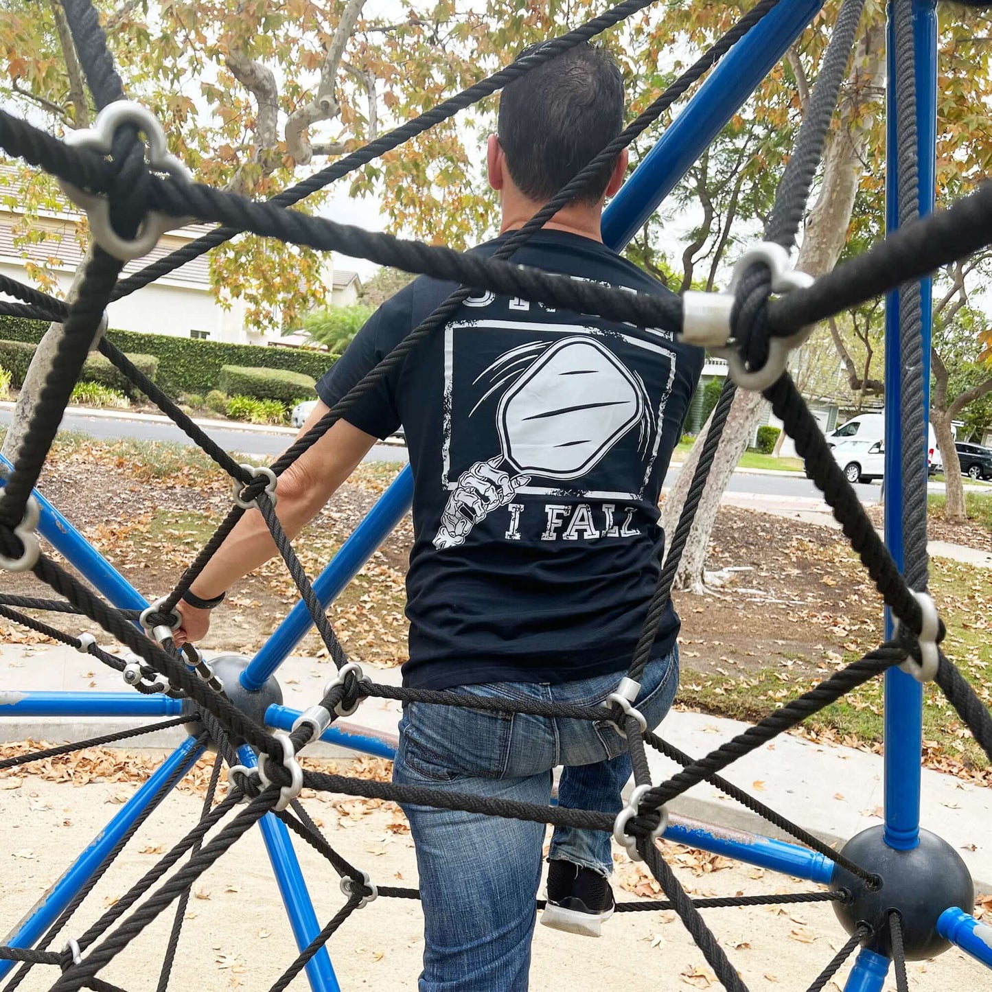 Man wearing "Until I Fall" Pickleball black t-shirt climbing jungle gym, showcasing motivational sportswear design.