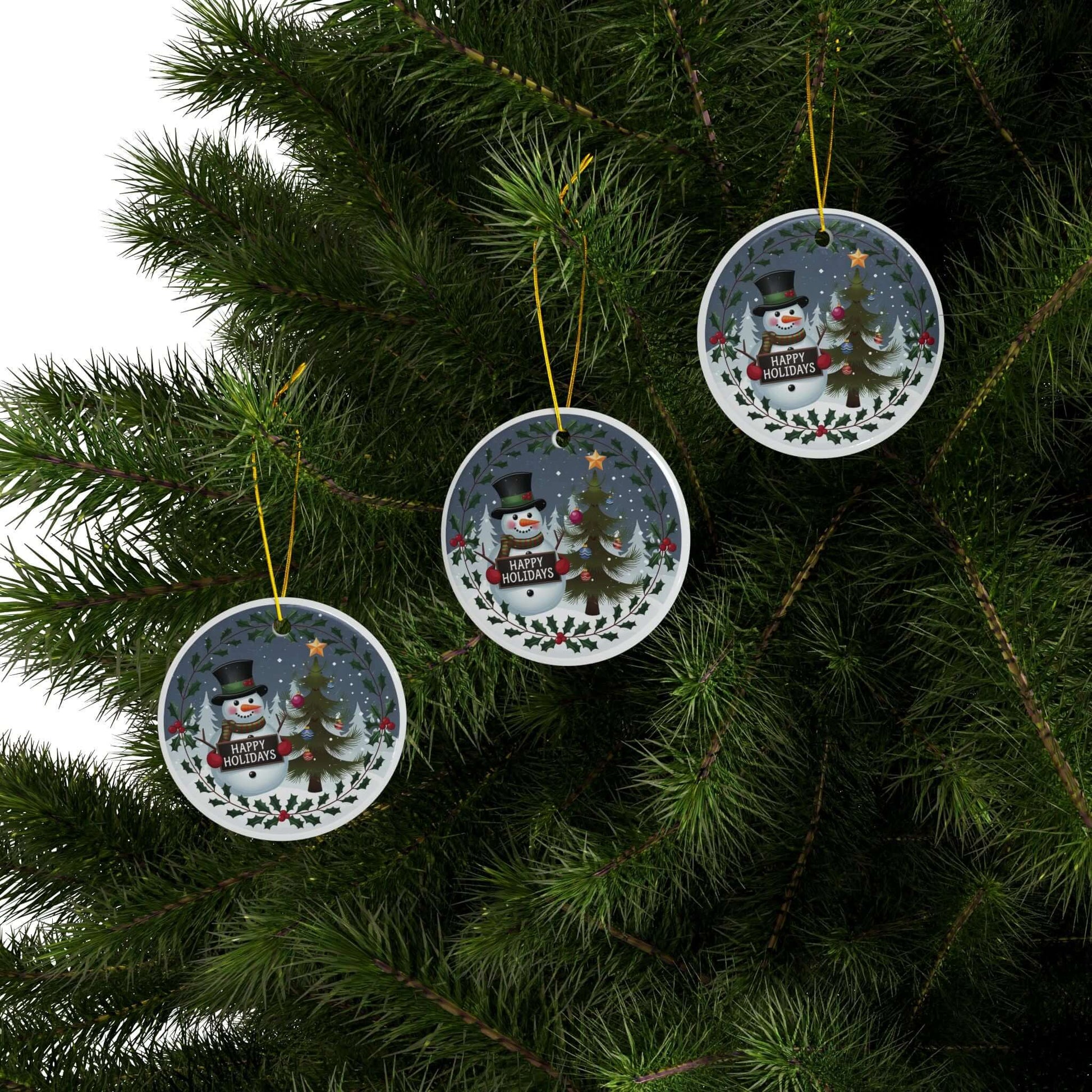 Festive two-sided ceramic ornaments featuring jolly snowman and Christmas tree hang on a lush green tree branch.