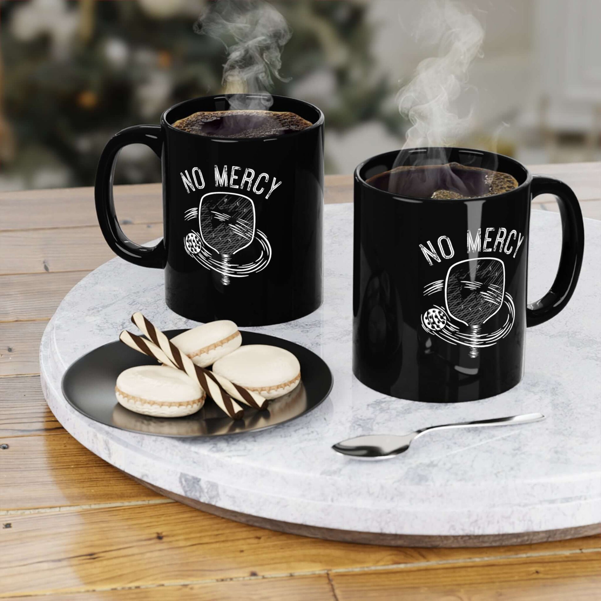 Two black "No Mercy" ceramic mugs with steam on a table, accompanied by a plate of macarons and cookies.