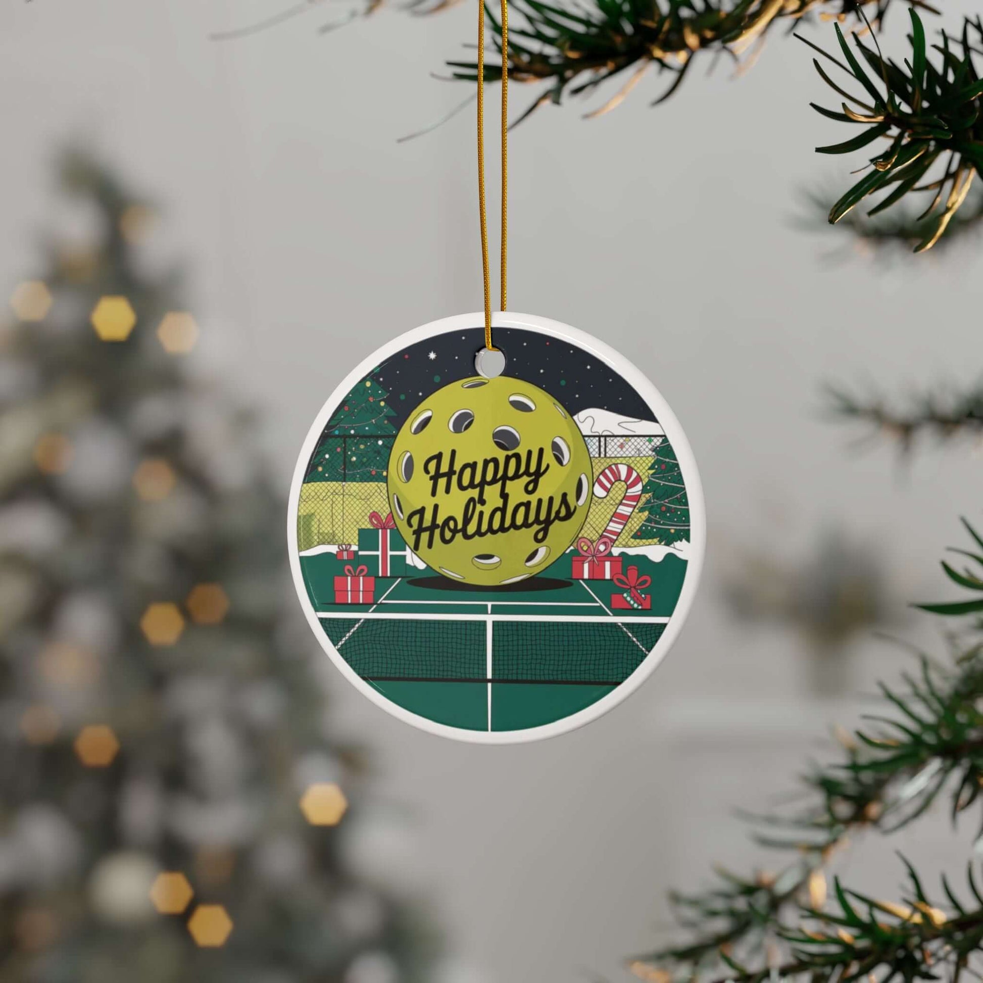 Festive pickleball holiday ornament with "Happy Holidays" text, Christmas tree, and gifts, hanging on a decorated tree branch.
