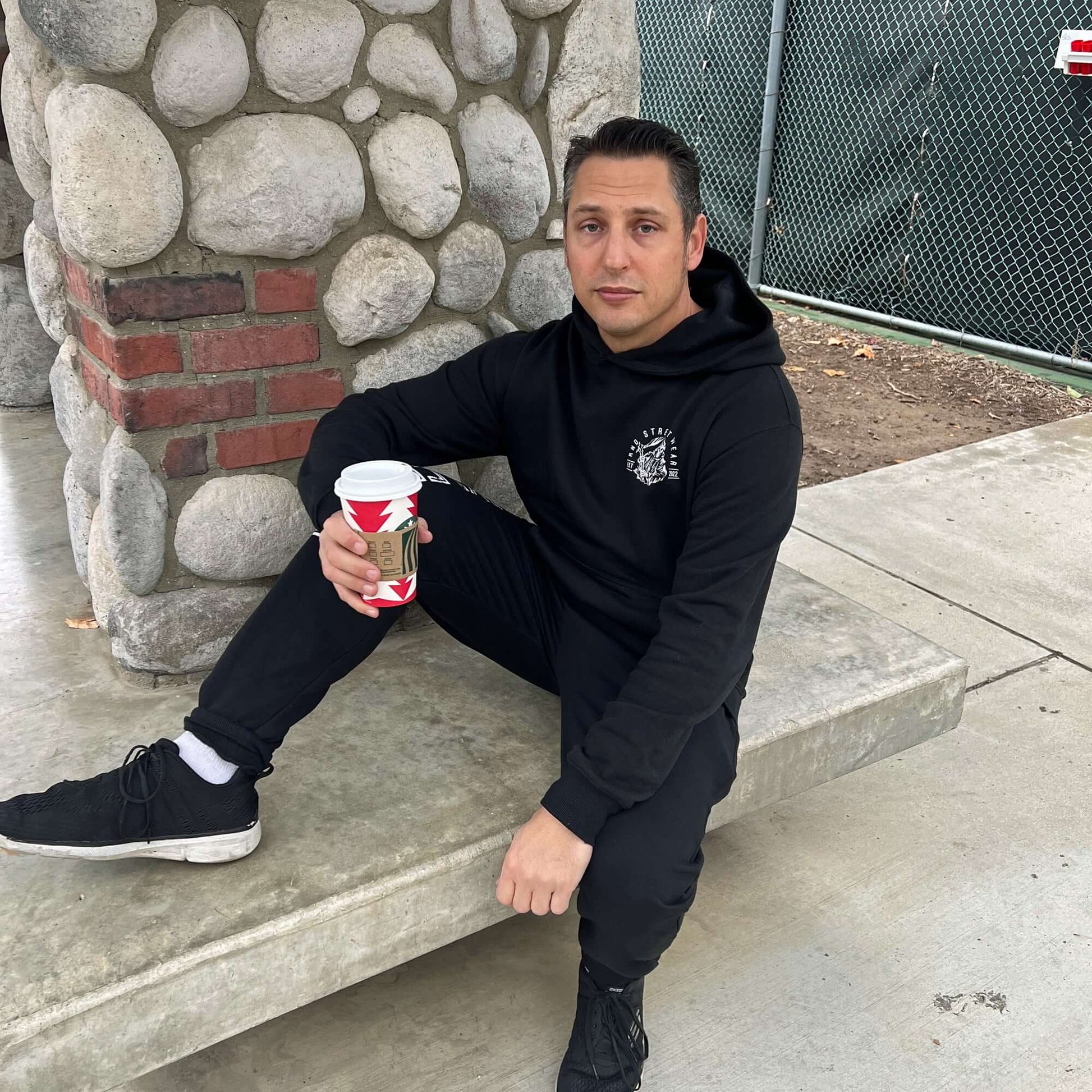 Man sitting on bench wearing black joggers and hoodie, holding a cup of coffee.