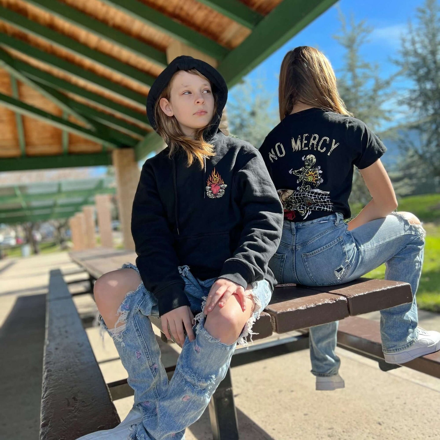 Two people wearing No Mercy apparel with skeleton and flaming heart design, sitting on a bench under a wooden canopy.