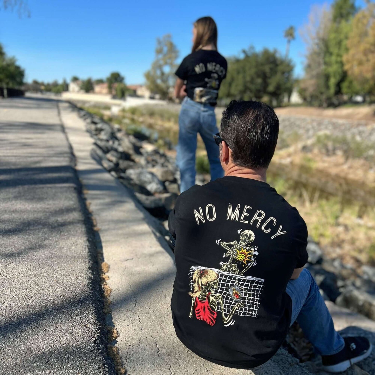 Two people wearing No Mercy Mixed Doubles Pickleball T-Shirts featuring skeletons with flaming hearts, outdoor setting.
