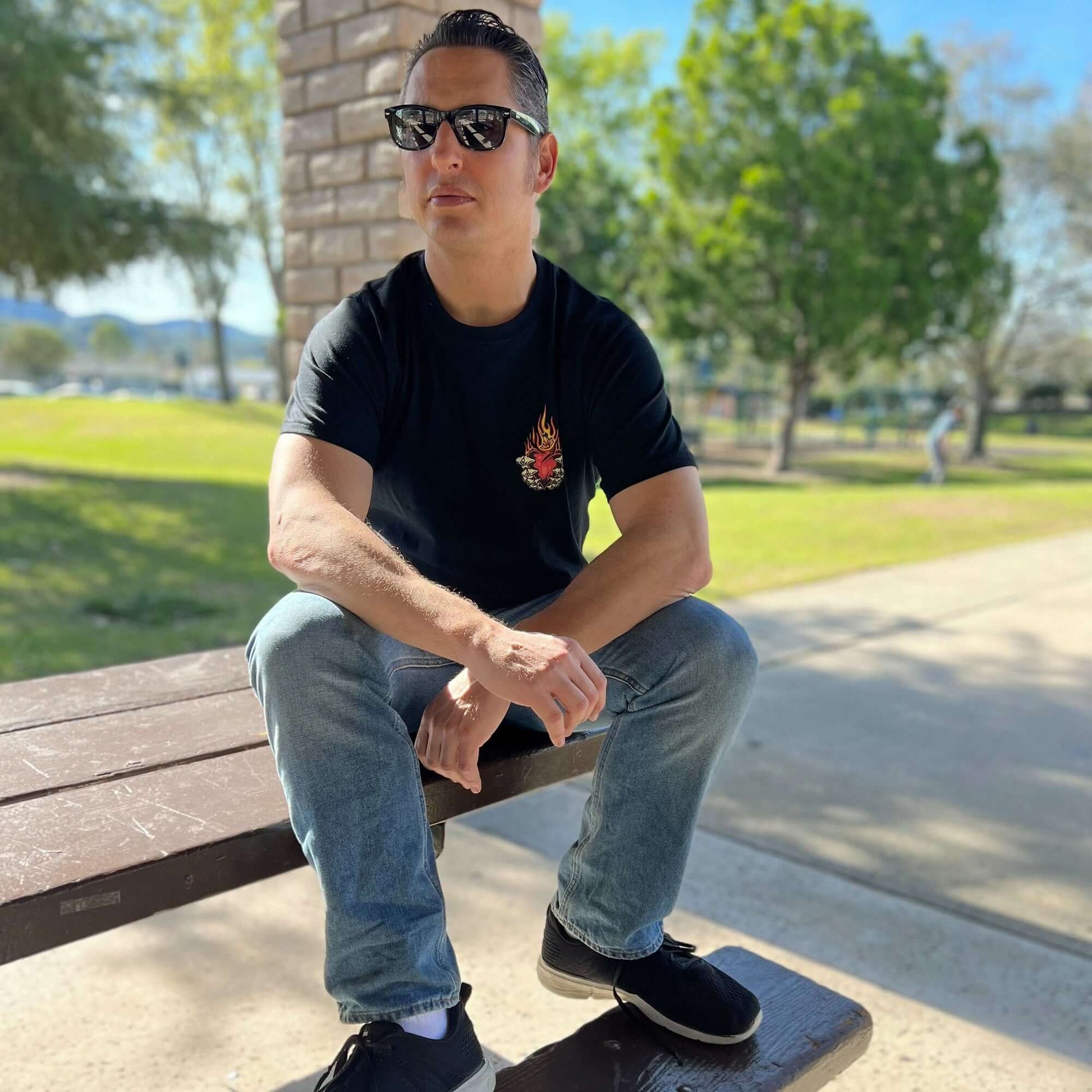 Man wearing No Mercy Pickleball T-shirt with flaming heart design sits on a park bench in sunglasses.