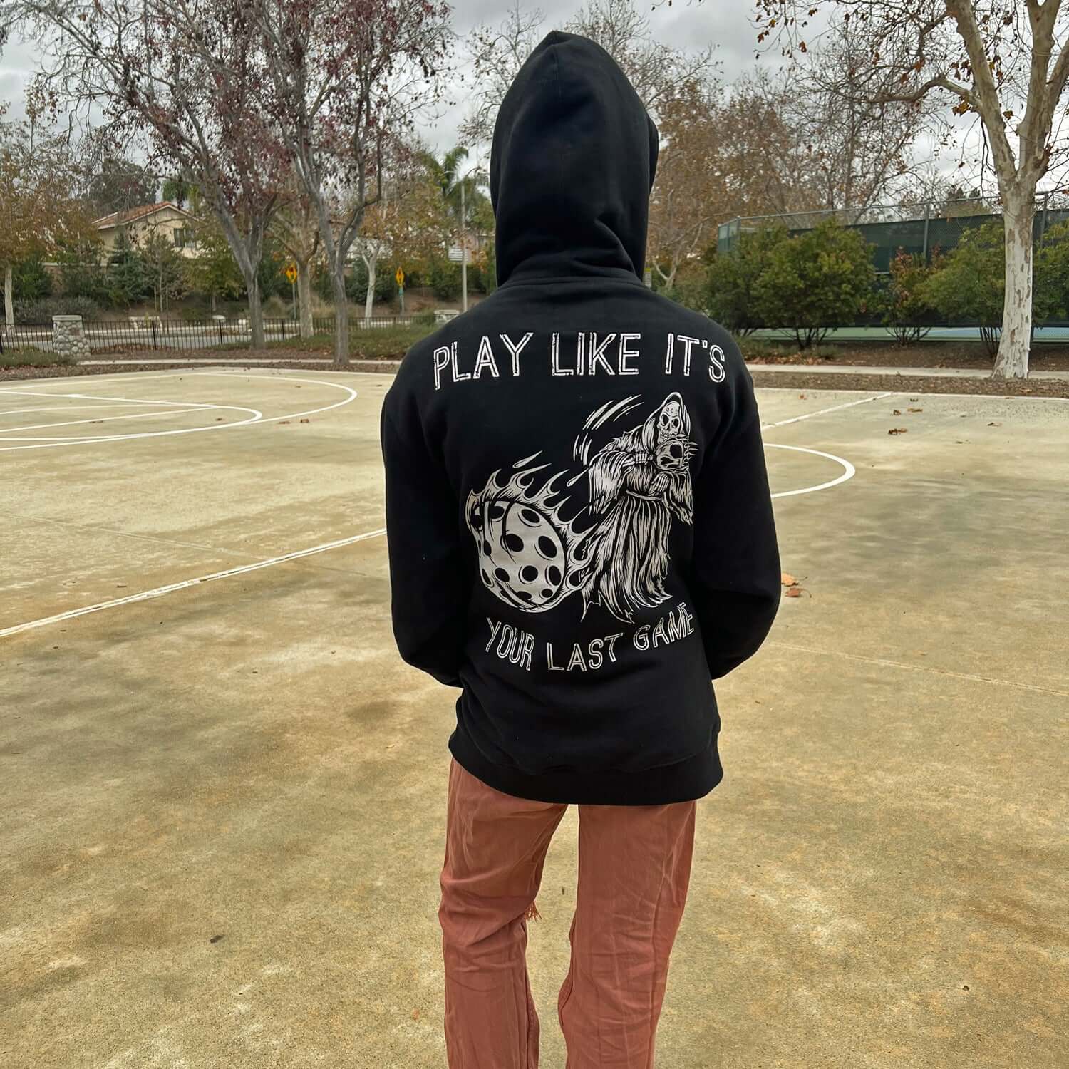 Person wearing black pickleball hoodie with "Play Like It's Your Last Game" and flaming reaper design on a tennis court.