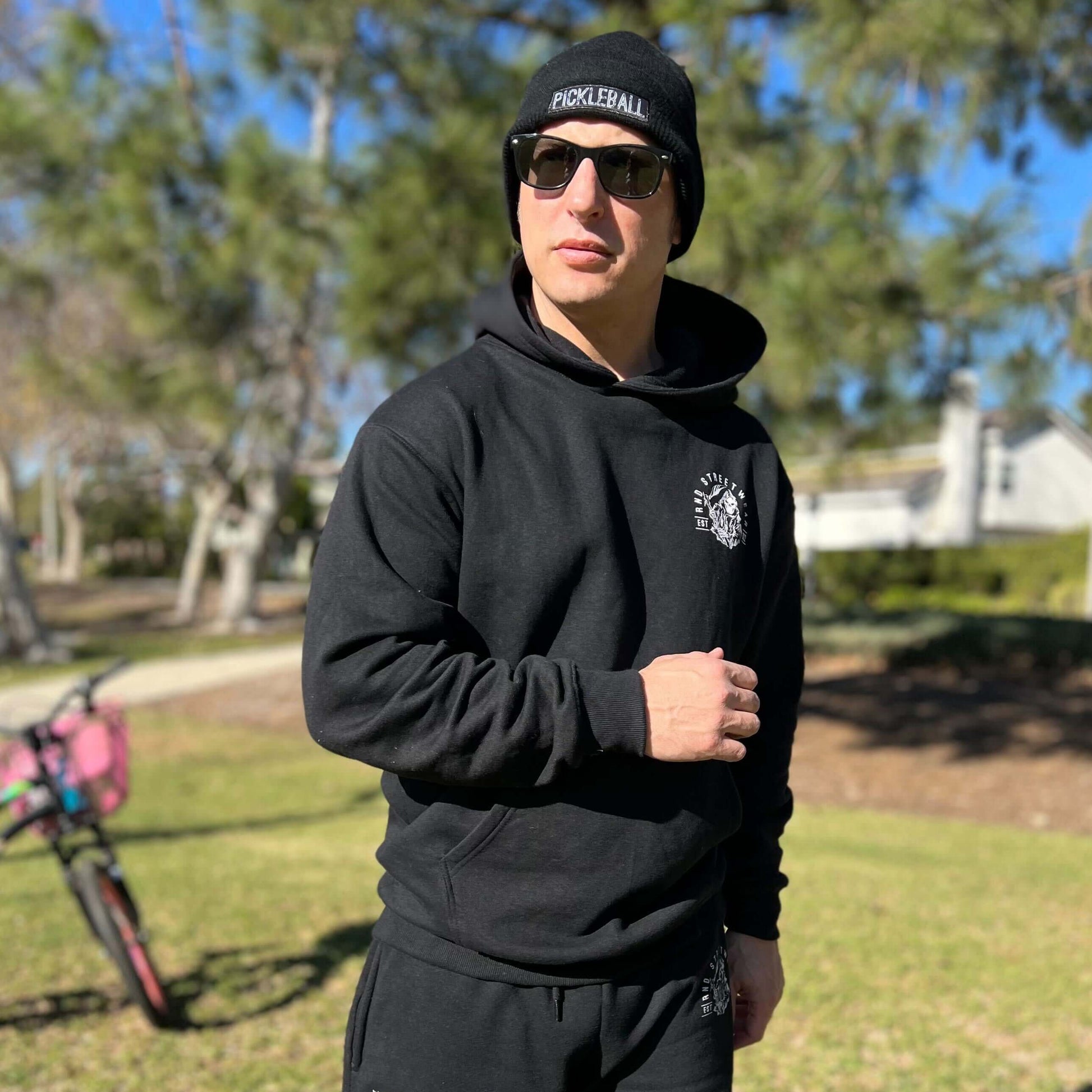 Man wearing a black pickleball hoodie with flaming reaper design, standing in a park with sunglasses and beanie.