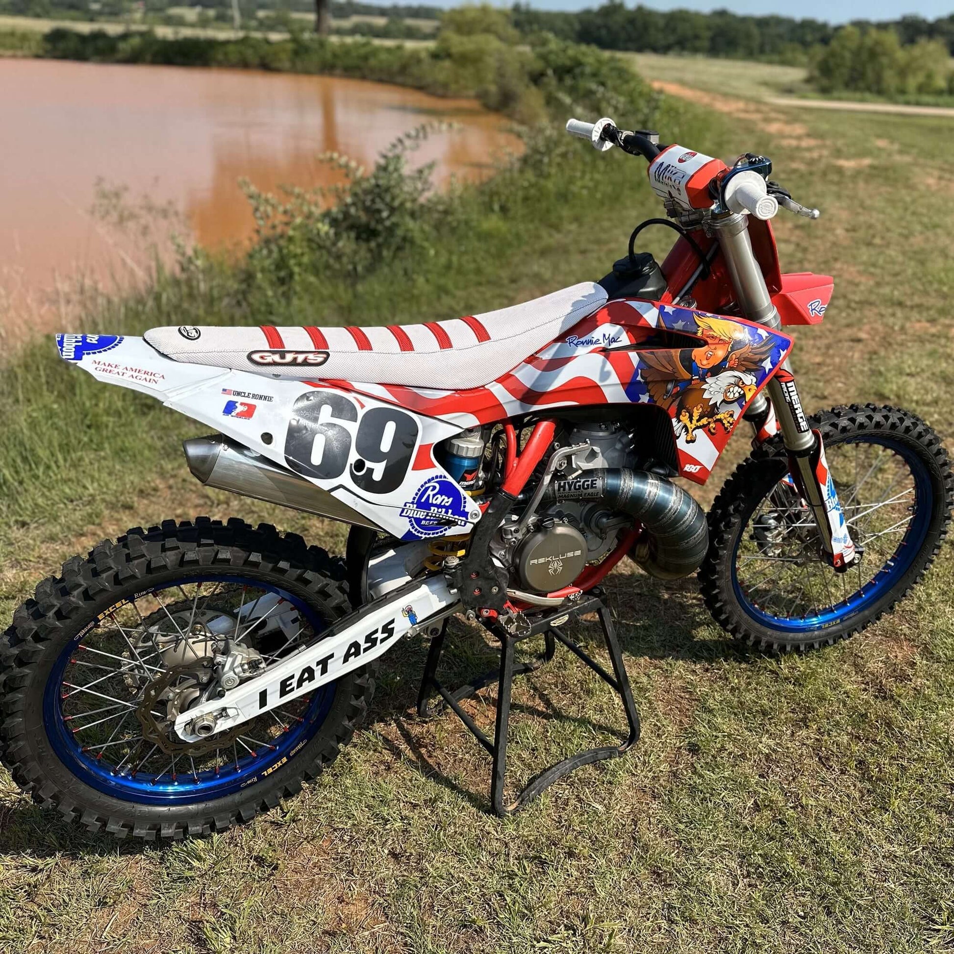 Red dirt bike with "I EAT ASS" swingarm decals, featuring American flag graphics and number 69, parked near a pond.